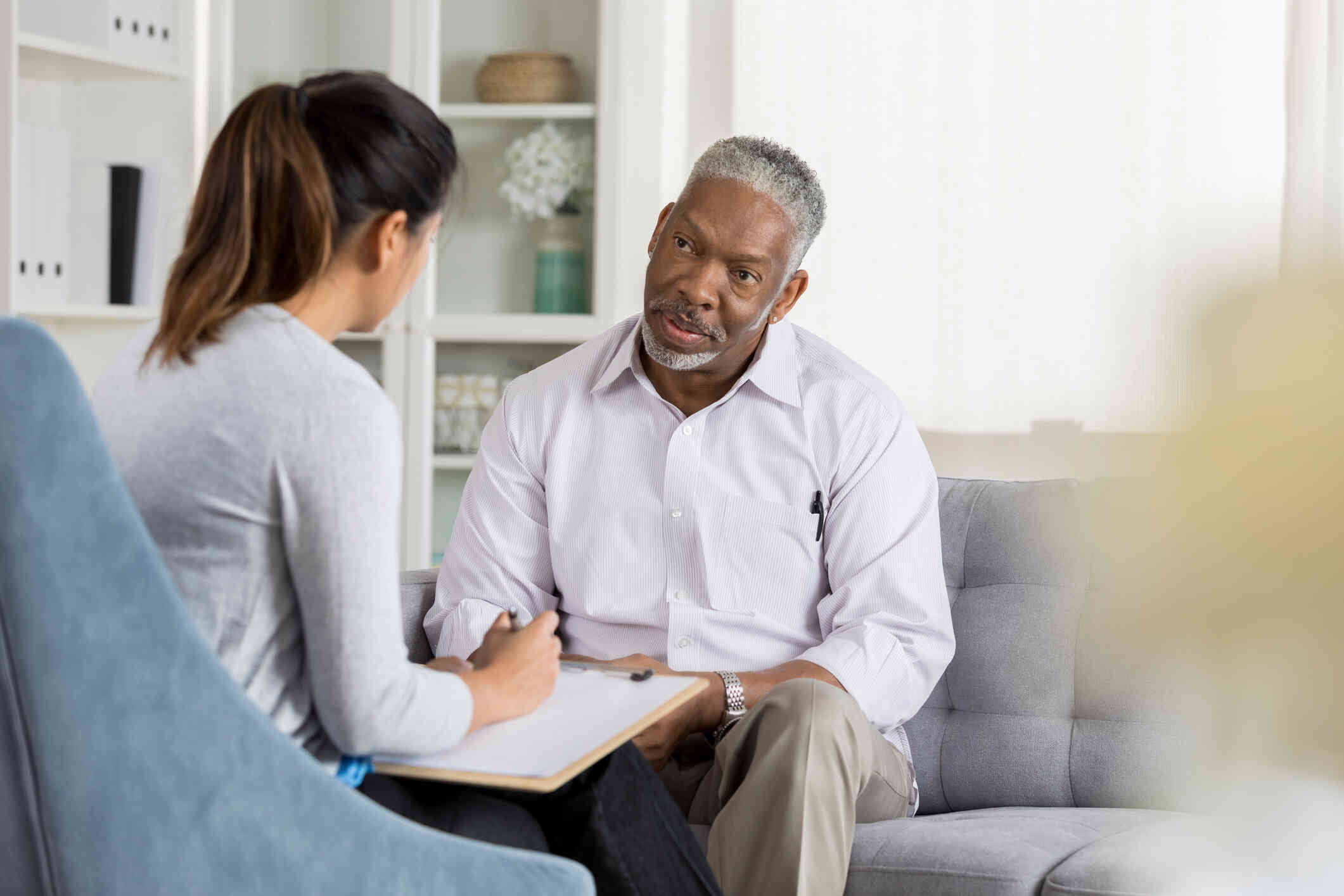 A middle aged man in a button down shirt sits on a couch and listens to the female therapist sitting across from him.